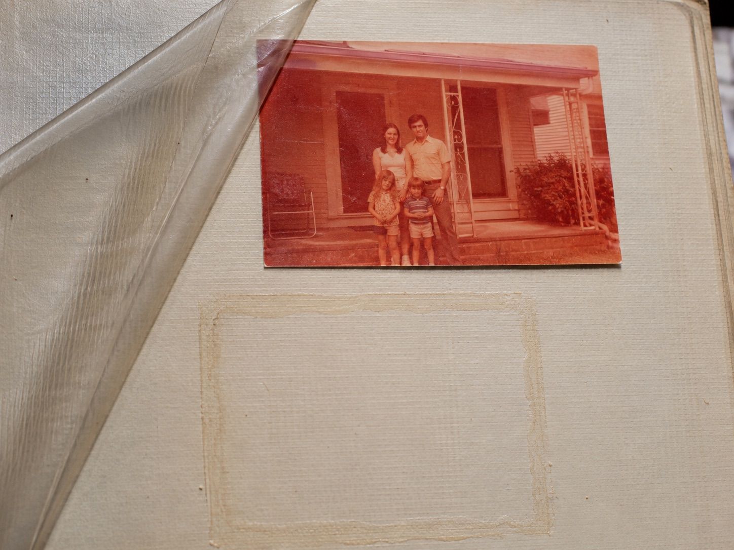 A 1974 color snapshot of a young family on a porch, faded to orange and salmon, stuck to the page of a magnetic photo album with the cloudy plastic sheet peeled back and a pale rectangle where another print was removed