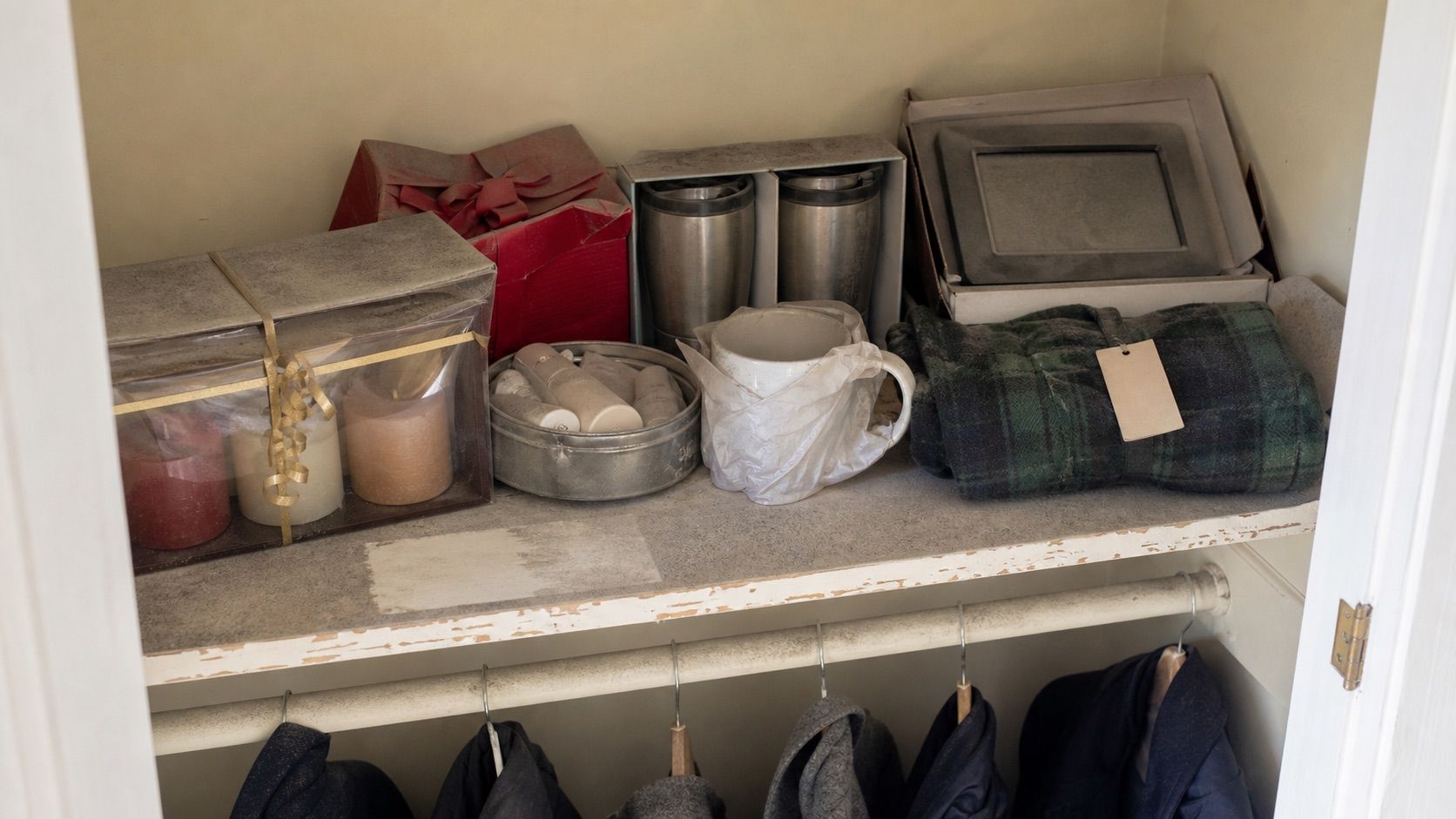 The top shelf of a hall closet, door open: a candle set still in cellophane, a lotion tin, two travel mugs in their boxes, a plaid fleece with the tag on and a boxed picture frame, all under a layer of dust, with coats hanging below