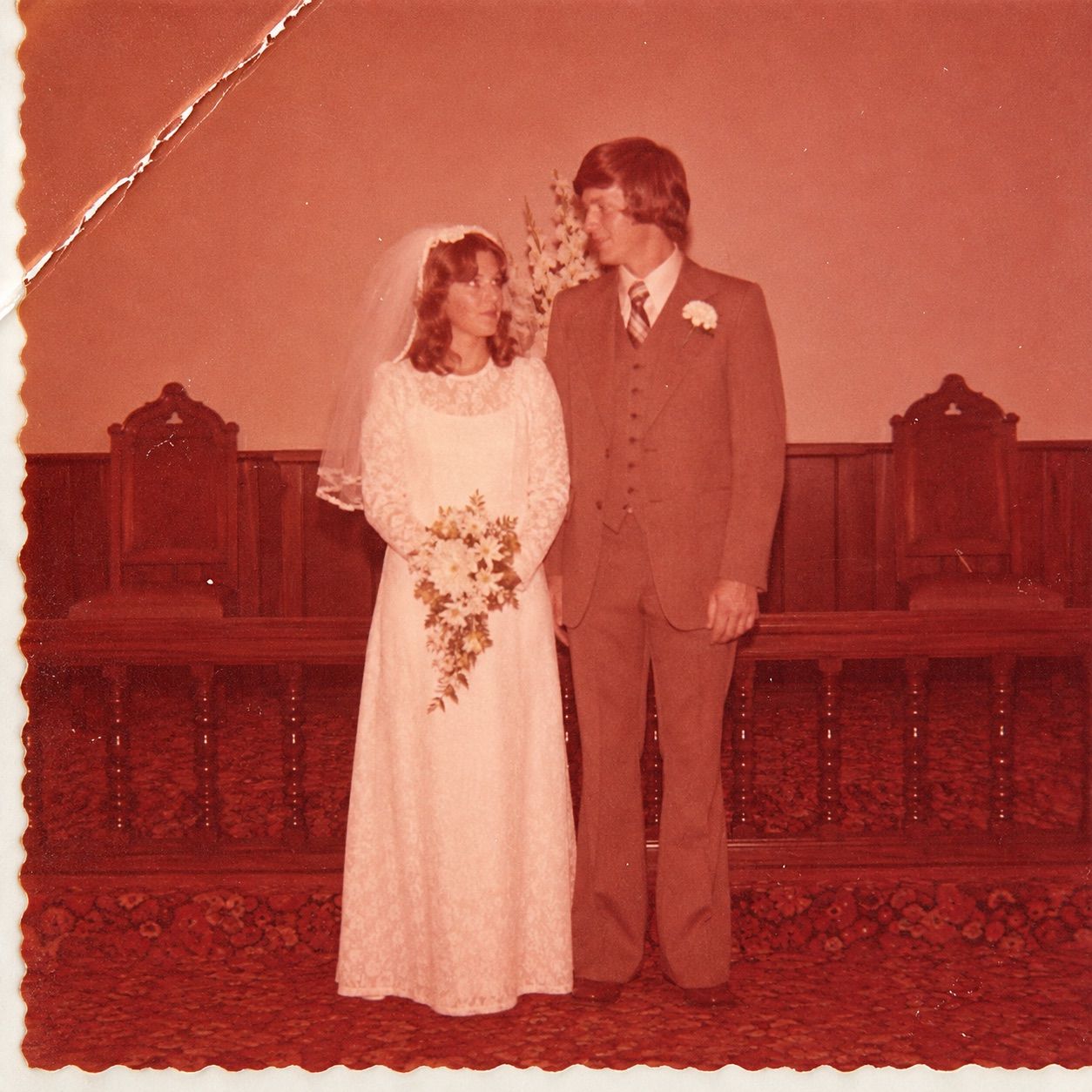 Scan of a 1976 church wedding print before restoration: heavy orange and magenta cast, milky shadows, a diagonal crease with a torn edge in the top left corner and white scratches across the bride's veil