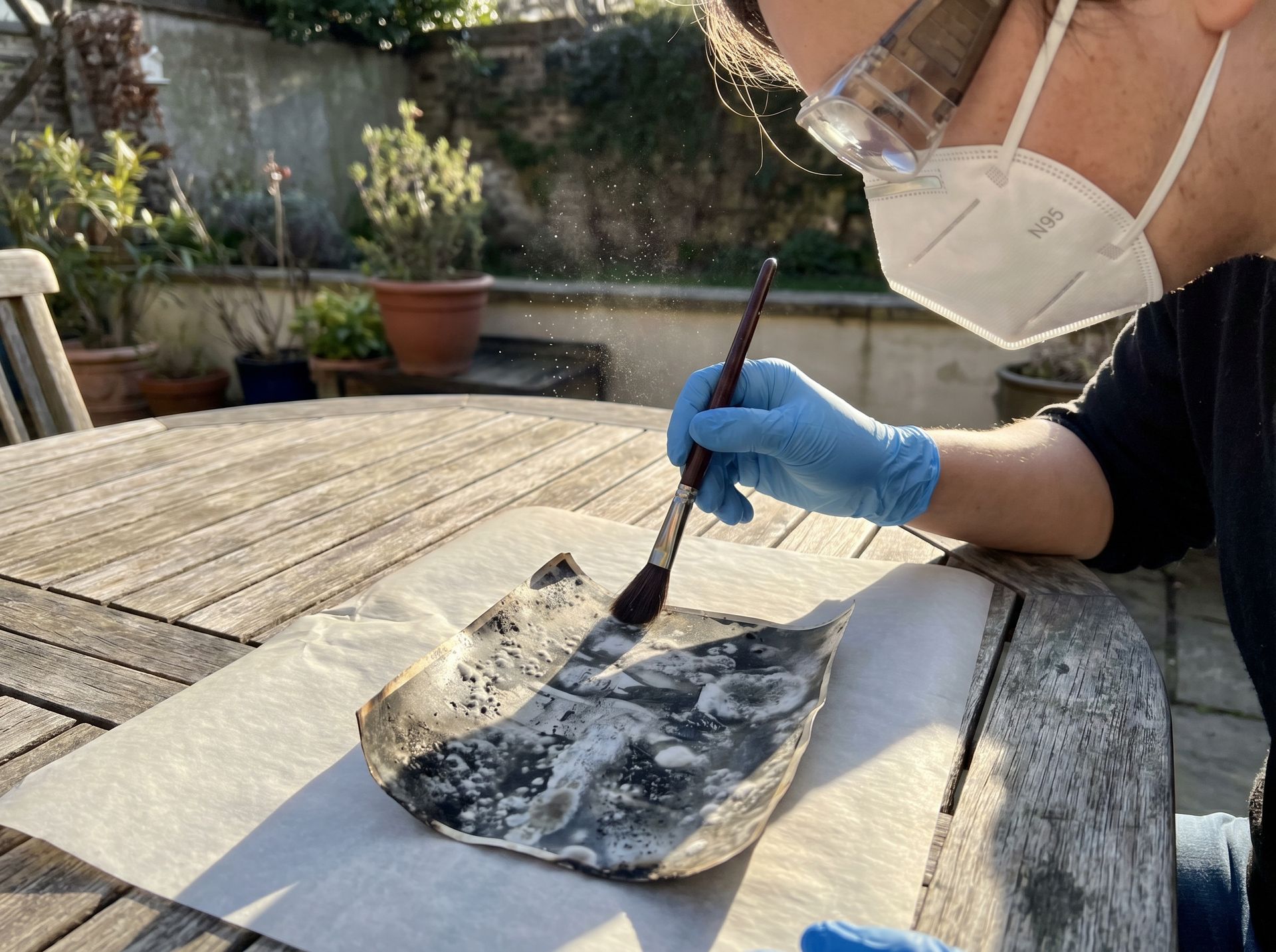 Person outdoors wearing N95 respirator and nitrile gloves carefully brushing a mold-damaged vintage photograph with a soft sable brush